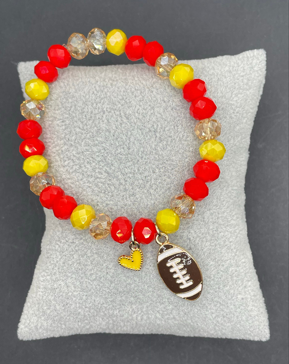 A beaded stretch bracelet with red, yellow, and clear faceted beads, featuring a brown football charm and a small yellow heart charm, displayed on a white cushion against a dark gray background.